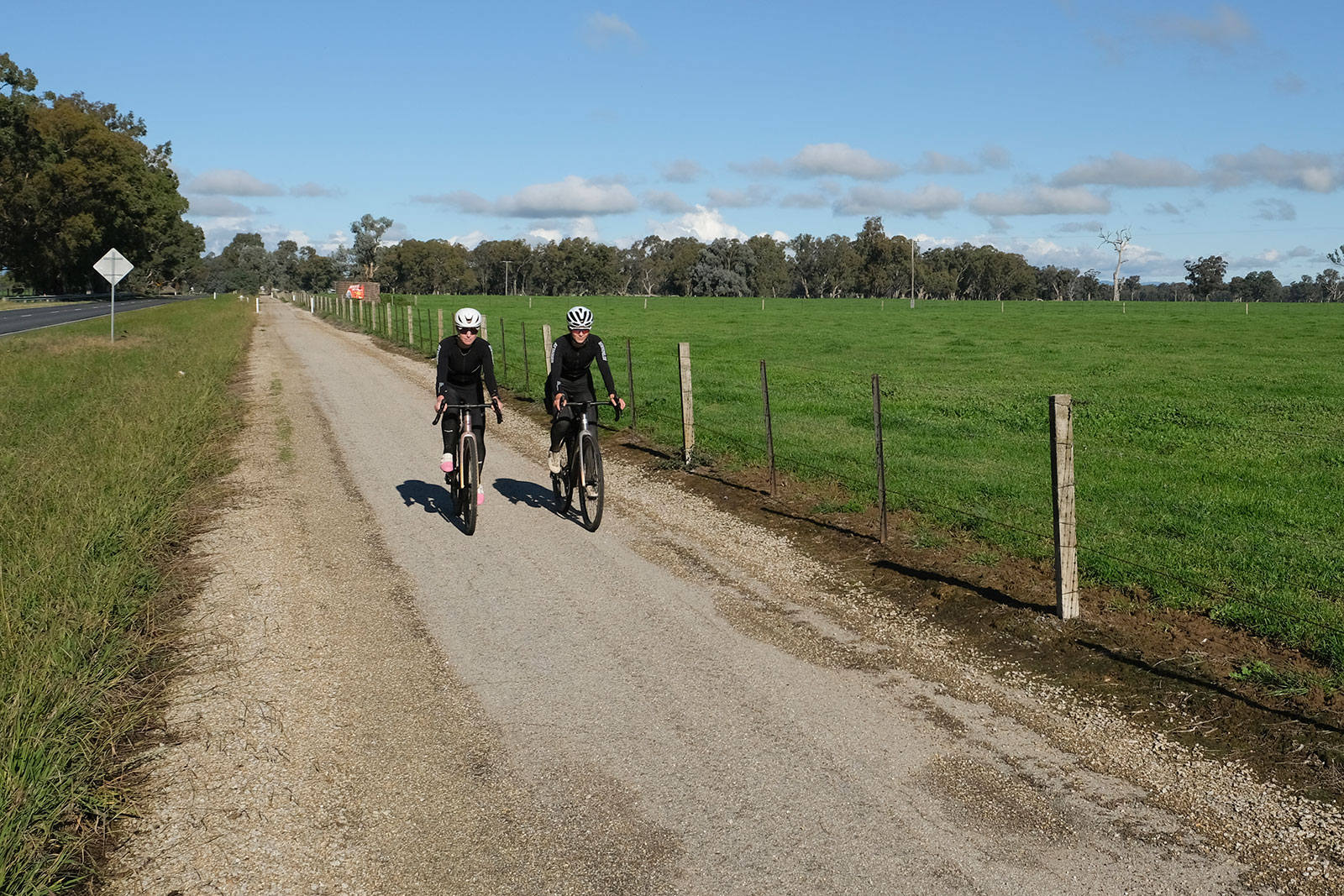 Two cyclists wearing helmets ride along a gravel country path beside a wooden fence and green fields under a bright blue sky.